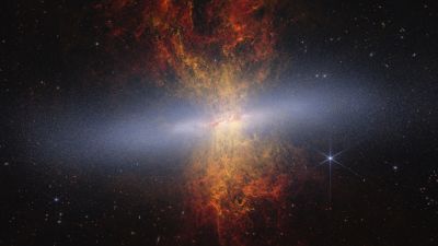 Webb and Hubble composite image of the edge-on starburst galaxy Messier 82, the Cigar Galaxy, showing millions of blue-white stars in the disk and orange plumes of dust and ionised gas streaming above and below it.